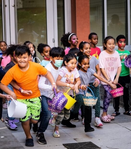 True-Ground-About-True-Ground-At-a-Glance A group of children, holding colorful baskets, excitedly participate in an outdoor event, likely an egg hunt or similar activity.