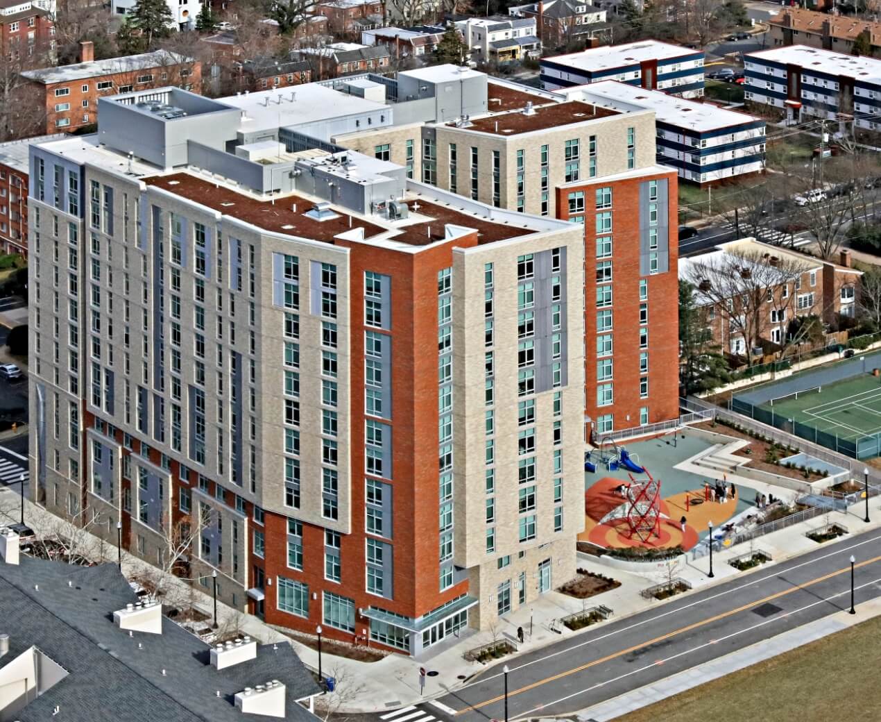 True-Ground-Home-Queens-Court-Aerial Aerial view of a modern multi-story residential building with a playground and adjacent street.