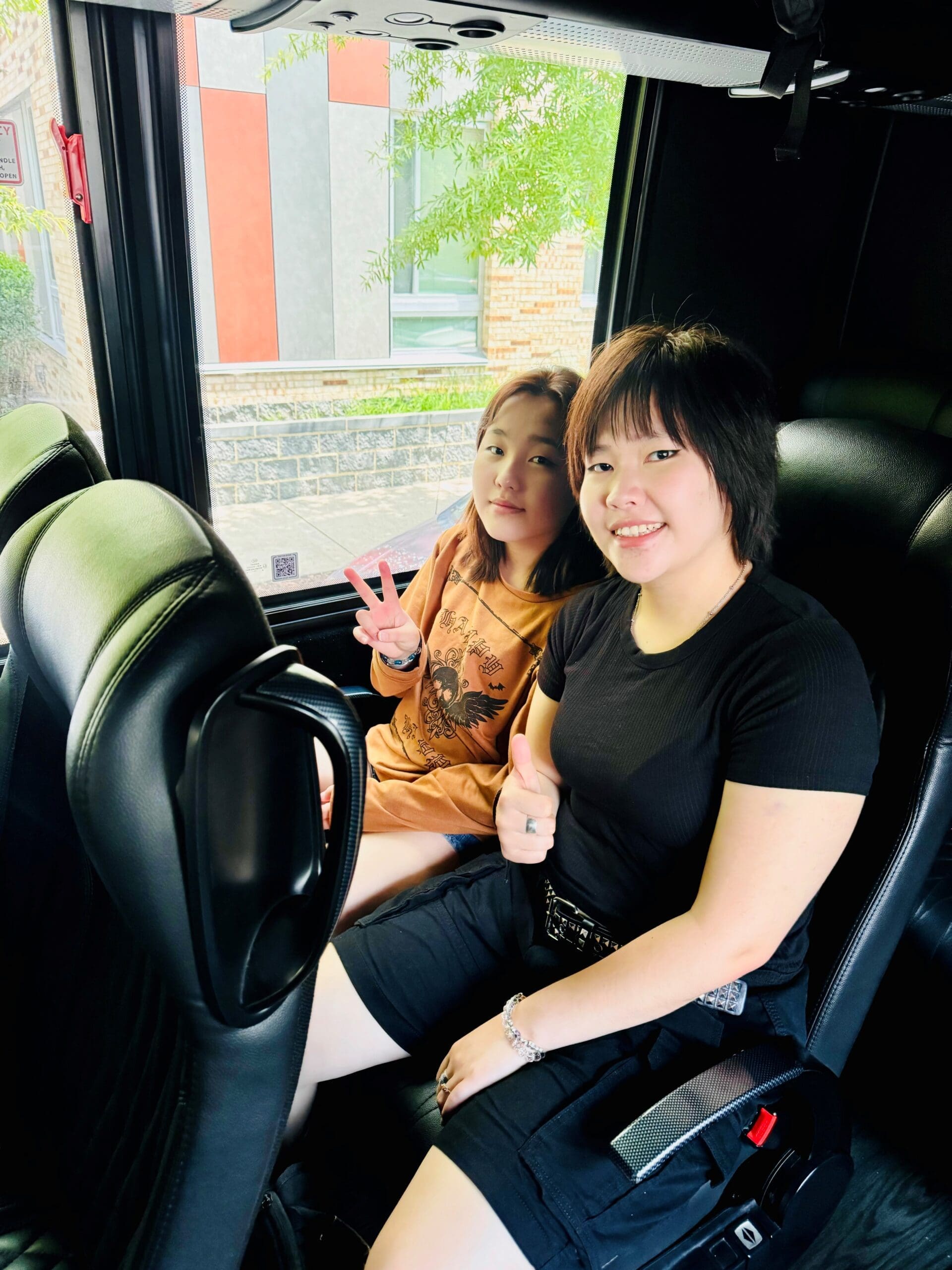 Two girls sitting on a bus with black seats, smiling for the photo.