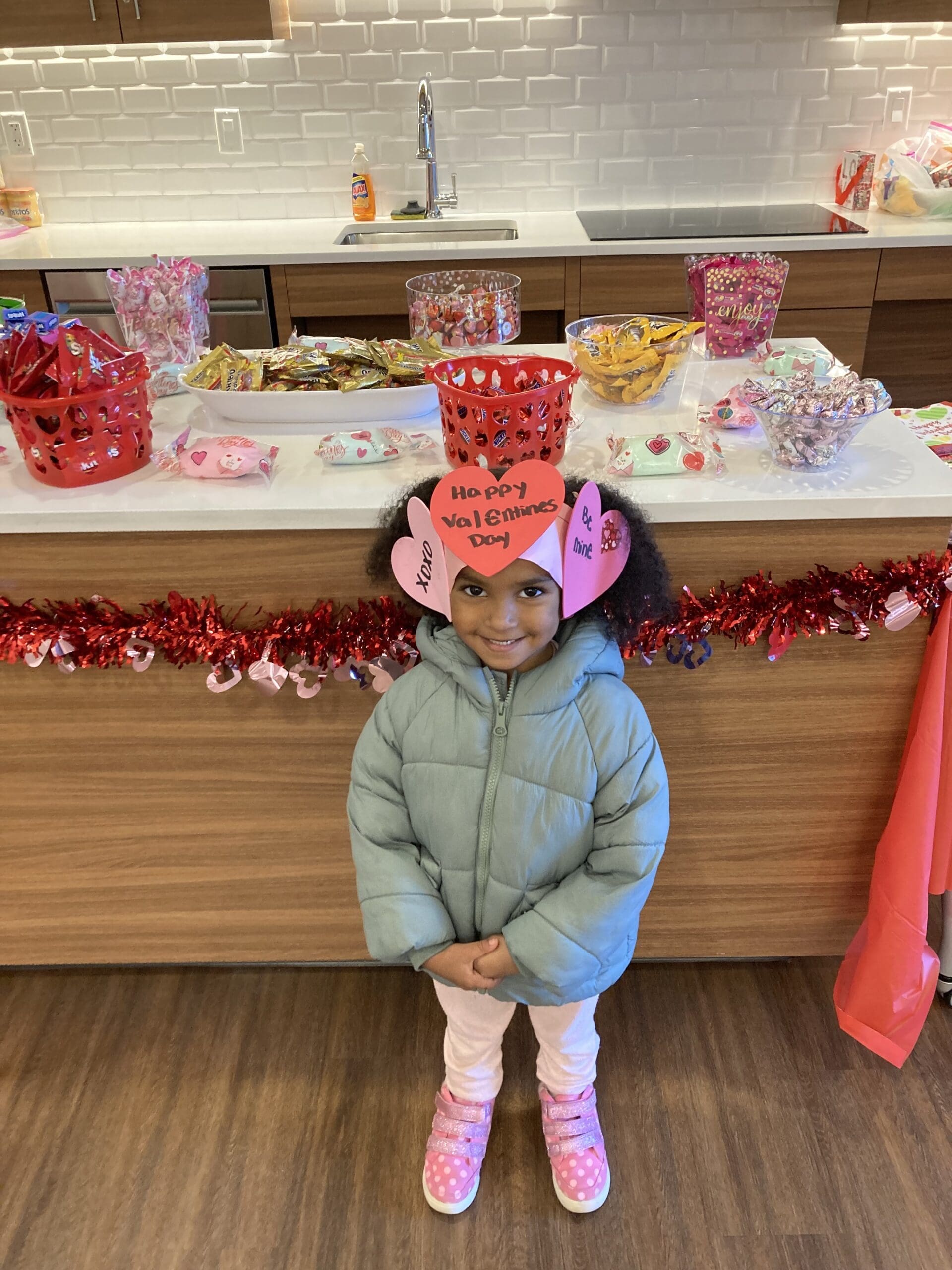 Girl smiling for the photo, she is in a kitchen, on her head you can see a decorative hair clip for valentine's day.