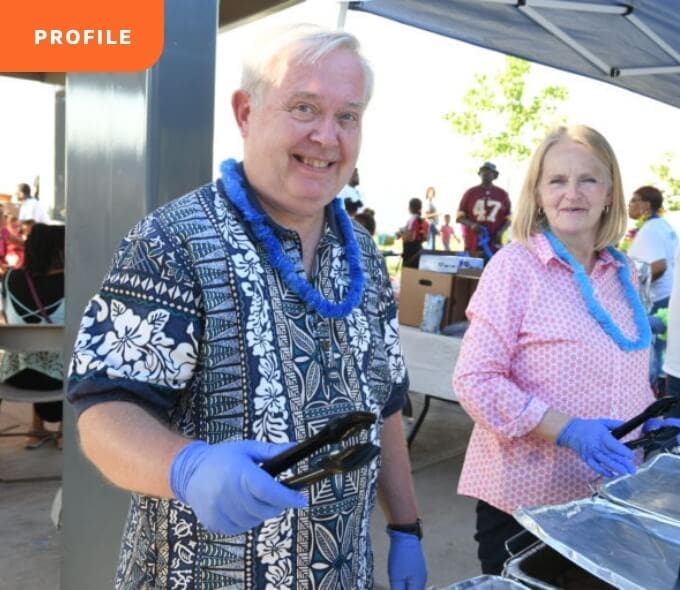 Impact-Stories_True-Ground-Meet-David@2x Two people in leis and gloves serve food at an outdoor event under a tent.