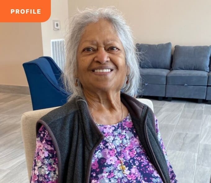 Smiling elderly woman with gray hair, floral top, dark vest, seated with gray couch in the background.