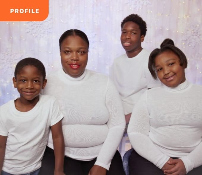 Impact-Stories_True-Ground-Meet-Jennifer@2x Family of four in white shirts, smiling in front of a snowy backdrop with twinkling lights.