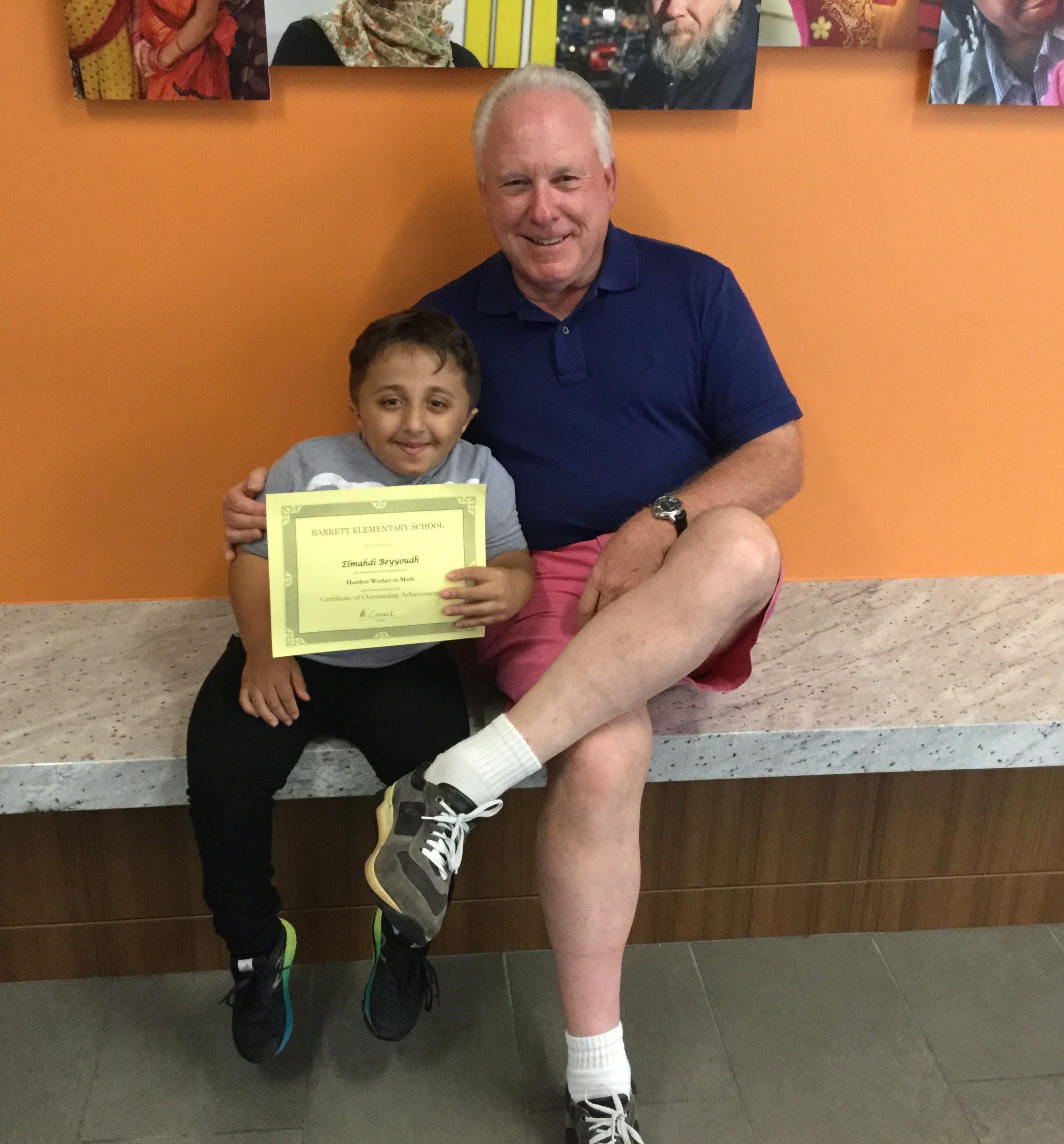 Child receiving a certificate, sitting with an adult in front of an orange wall