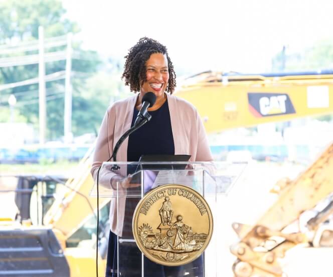 A woman stands at a podium with the seal of the District of Columbia, smiling as she speaks into a microphone, with construction equipment visible in the background.