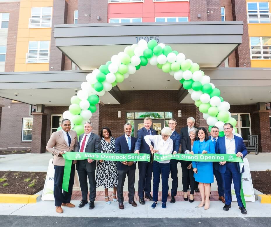 Group of people cutting a green ribbon at the grand opening of Ida's Overlook Senior Residences, under a balloon arch.