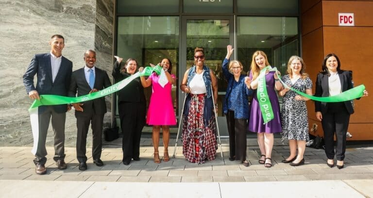 A group of people, stand outside a building, holding a green ribbon and celebrating a ribbon-cutting ceremony.