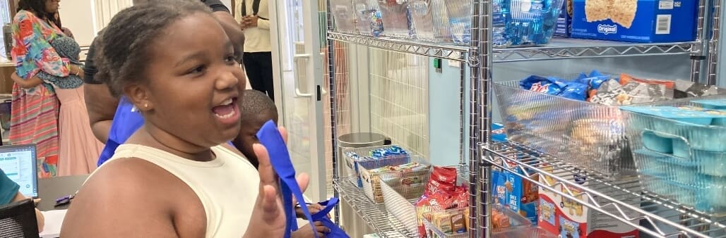 Girl smiling and holding a blue tote bag while selecting items at a community shelf