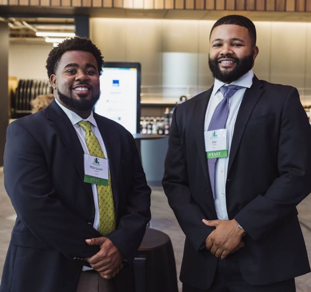 True-Ground-How-to-Help-Work@2x Two men in suits smiling at a professional event, wearing staff badges