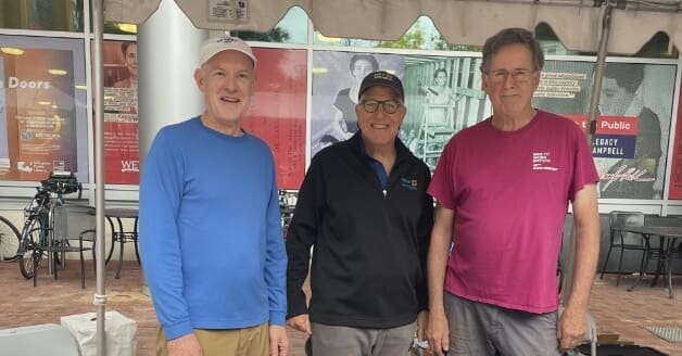 Three men in casual attire smiling under a tent at an outdoor event