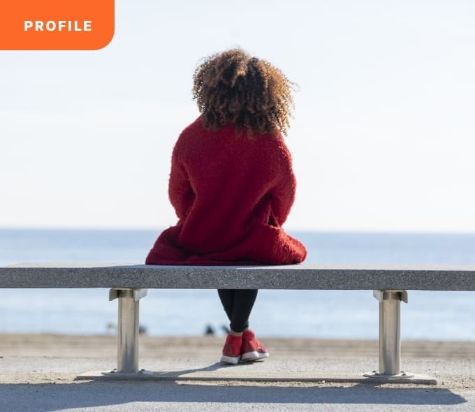 True-Ground-Meet-Harmony@2x Curly-haired person in red sweater on bench, facing ocean, "Profile" tag top left.