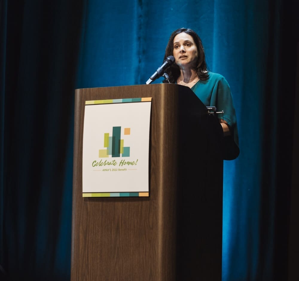 Woman in green dress speaking at a podium with a "Celebrate Home!" sign.